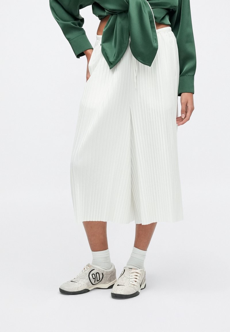Personne portant un culotte plissé blanc, des chaussettes blanches, des baskets blanches à motifs avec le numéro 90 et un haut vert à manches longues noué.