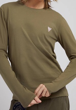 Personne portant une chemise à manches longues vert olive avec un petit logo triangulaire « Guess » sur la poitrine et un pantalon assorti vert olive, sur un fond uni.