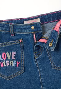 Jeans de mezclilla con "LOVE THERAPY" bordado en letras coloridas. Presentan una cremallera rosa y costuras del mismo color, además de un detalle de corazón en el bolsillo trasero.
