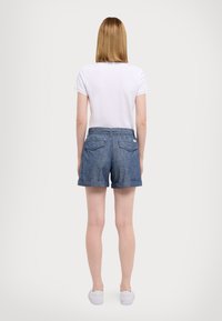 Person med rakt blont hår som bär en vit t-shirt, blå shorts med upprullade kanter och bakfickor, samt vita sneakers, stående med ryggen vänd.