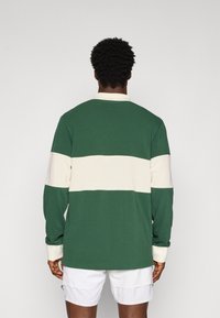 Longsleeve polo w ciemnozielonym kolorze, z białymi poziomymi paskami. Materiał ma miękką teksturę. Posiada klasyczny kołnierzyk i ściągacze na rękawach.