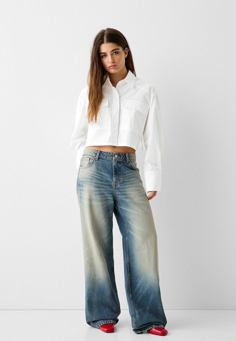 Camicia bianca corta con maniche lunghe e due tasche sul petto, abbinata a jeans azzurri svasati e scarpe basse rosse.