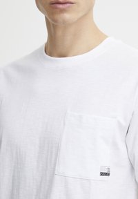White cotton t-shirt featuring short sleeves, a round neck, and a left chest pocket with a small logo label. Soft texture, simple design.