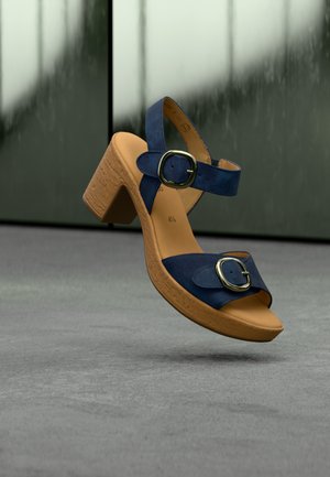 Blauer Wildleder-Pumps mit hohem Blockabsatz, breitem Vorderriemen und zwei runden Metall-Schnallen. Beigefarbene profilierte Außensohle mit abgerundeter Zehenpartie.