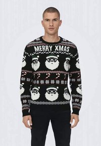Mann iført svart julesweater med gjentagende nisseansikter, teksten "MERRY XMAS" og pepperkake-mønstre.