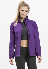 Lilla softshell-jakke med høj krave, lynlås lukning og sidelommer. Glat tekstur og tætsiddende design, parret med en sort top og leggings.