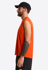 Camiseta sin mangas naranja hecha de tela ligera, con escote redondo. Se lleva con pantalones cortos negros y una gorra negra, perfil lateral visible.