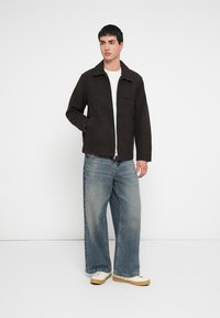 Dunkelbraune Jacke aus Wollmischung mit geradem Schnitt, einer Brusttasche und Seitentaschen, kombiniert mit weiten blauen Jeans und weißen Schuhen.