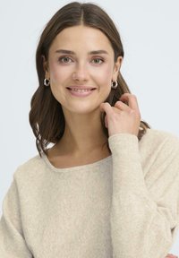 Ljusbeige tröja med rund halsringning och långa ärmar, tillverkad av mjuk, texturerad tyg. Modellen bär silvriga hoops och ler.