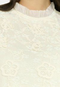 Top in pizzo bianco con ricami floreali, texture trasparente e scollatura a volant. Le cuciture dettagliate aggiungono profondità al design.