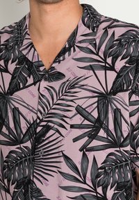 Koszula z krótkim rękawem z lekkiego materiału, z różowym tłem oraz dużym czarnym nadrukiem liści palmowych i fioletowymi akcentami tekstowymi.