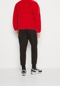 Chaqueta de forro polar roja con un acabado texturizado, pantalones de chándal negros con bolsillos laterales y zapatillas de correr negras con estampados blancos.
