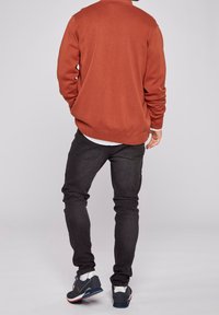 Röd ribbad tröja, svarta slim-fit jeans och blåa sportskor. Tröjan har en rund hals och stickad textur, designad med en avslappnad stil.