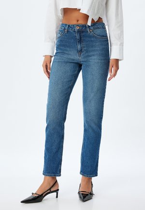 Jeans en denim bleu avec une coupe droite, présentant une taille haute, cinq poches et une texture légèrement décolorée, associés à des chaussures noires à petit talon.