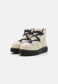 Beige høyhælte støvler med svarte gummisåler, teksturert ulloverdel og krysslagte svarte lisser. Siden har en glatt imitert skinnfinish.