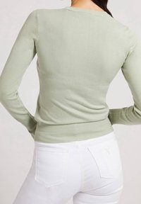 Haut en tricot vert clair à manches longues avec un design ajusté, un col rond, des poignets côtelés et une ceinture. Porté avec un jean blanc.