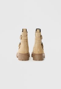 Bottines en suède beige avec une épaisse semelle en caoutchouc marron, des sangles latérales ajustables et une fermeture éclair à l'arrière. Surface texturée pour un meilleur grip.