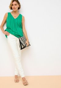 Blusa verde senza maniche con scollatura a V e parte anteriore plissettata. Abbinata a pantaloni skinny bianchi e una borsa a tracolla a righe bianche e nere. Infradito.