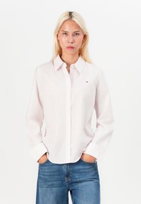 Camicia rosa chiaro a maniche lunghe con righe verticali, colletto a camicia, bottoni bianchi e logo ricamato sul petto sinistro. Abbinata a jeans blu.