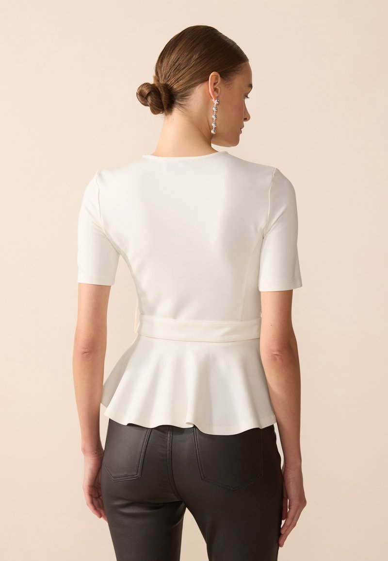 Weißes, eng anliegendes Oberteil mit kurzen Ärmeln, schwingendem Peplum-Saum und Bindeband im Rücken. Trägt dazu hochtaillierte schwarze Hose und silberne Ohrhänger.
