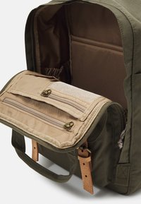 Olivengrønn ryggsekk med brune detaljer, med glidelåser, en nettlomme og et solid håndtak; innvendig foret med brunt stoff.