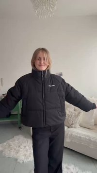 Black puffer jacket with a high collar and front zipper, featuring a logo on the left chest. It has a boxy fit and soft texture.