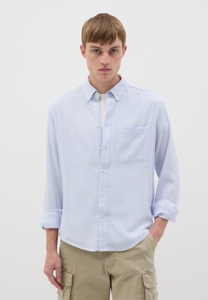 Light blue button-up shirt with long sleeves, chest pocket, and roll-up cuffs, made from a textured fabric. Paired with khaki cargo shorts.