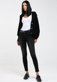 Cardigan negro de punto con grandes botones, llevado sobre una camiseta blanca sin mangas. Combinado con jeans negros y zapatos tipo slip-on negros. Texturas suaves.
