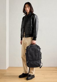 Black leather bomber jacket with a zip front, beige trousers, and a black fabric backpack with multiple zip compartments. Smooth texture.