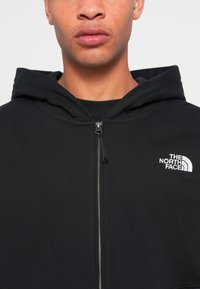 Black zip-up hoodie made of ribbed fabric. Features a drawstring hood and embroidered white logo on left chest. Metal zipper detail.