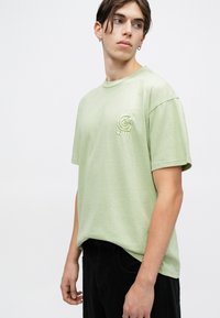Camiseta de algodón verde claro con un corte relajado, mangas cortas y un pequeño logo bordado en el lado izquierdo del pecho. Pantalones negros completan el conjunto.