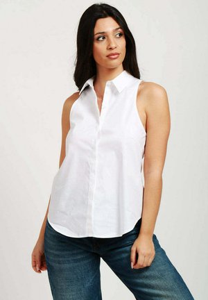 Blusa bianca senza maniche con collo a camicia, apertura frontale con bottoni e vestibilità ampia, abbinata a jeans in denim blu. Texture liscia.