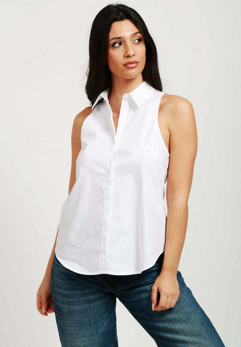 Blusa bianca senza maniche con collo a camicia, apertura frontale con bottoni e vestibilità ampia, abbinata a jeans in denim blu. Texture liscia.