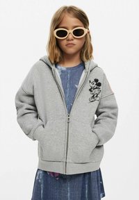 Szürke, cipzáros kapucnis pulóver, puha anyagból készült, Mickey Mouse mintával a mellrészen és bordázott szegéllyel. Farmerruhán viselve.