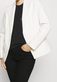 Vit blazer med skräddarsydd passform, enfaldig knäppning, snedskurna kragar och slät textur, bärs över en svart topp och byxor.