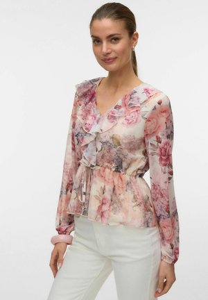 Femme portant un chemisier floral transparent à volants et manches longues, associé à un pantalon blanc, souriante avec les cheveux attachés.