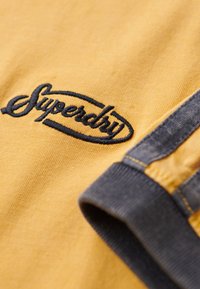 Gele katoenen t-shirt met marineblauwe geborduurde "Superdry" logo en grijs ribbenmouw. Heeft een gladde textuur en contrasterende kleurdetails.