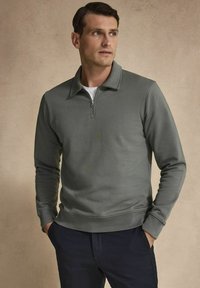 Grå stickad tröja med kvartslång dragkedja i kragen, långa ärmar och en mjuk yta, kombinerad med mörka byxor. Enkel, modern design.