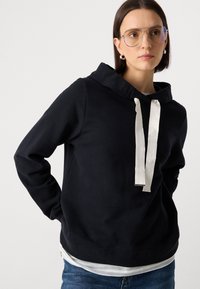 Tamsiai mėlyna gobtuvinė palaidinė, pagaminta iš švelnios medžiagos, su dideliu gobtuvu ir baltais raišteliais, papuošta įspaustu logotipu, derinama su šviesiai mėlynomis džinsinėmis kelnėmis.