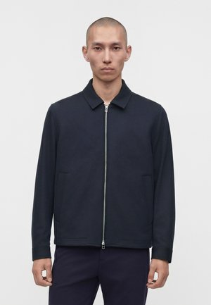 Hombre con la cabeza rapada, vestido con una chaqueta azul marino con cremallera y pantalones oscuros, de pie frente a un fondo liso de color gris claro.