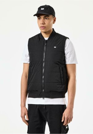 Sort polstret vest med lynlås foran, ribbet nederkant og sidelommer, båret over en hvid t-shirt. Sammen med en matchende sort kasket.