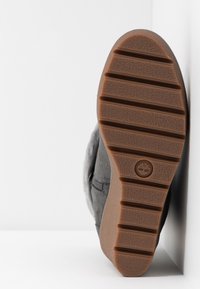 Semelle cannelée marron d'une botte grise reposant contre un mur blanc, montrant le logo Timberland centré sur la semelle.