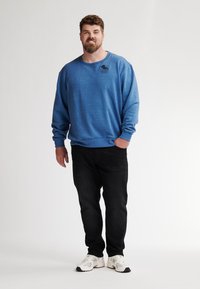 Sudadera azul de manga larga con cuello redondo y detalle gráfico negro, combinada con jeans negros y zapatillas deportivas blancas.