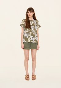Blusa a stampa floreale verde con maniche corte, abbinata a pantaloni shorts olive con orlo arrotolato. L'outfit presenta una vestibilità rilassata e un tessuto leggero.