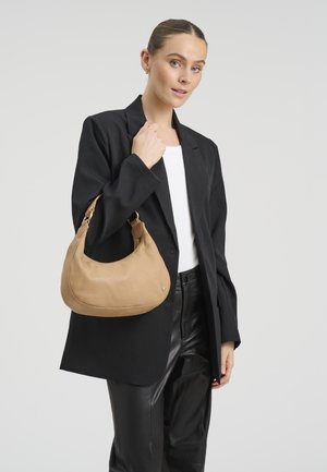 Mlada žena u crnom blejzeru i kožnim hlačama drži smeđu kožnu torbu preko ramena na bijeloj pozadini.