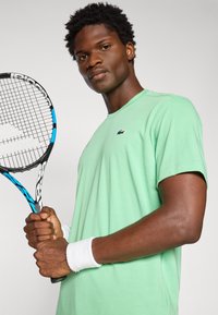 Limetkovozelené bavlnené tričko s okrúhlym výstrihom a krátkymi rukávmi, na ktorom sa nachádza malé logo Lacoste. Osoba drží tenisovú raketu s čiernymi a modrými akcentmi.