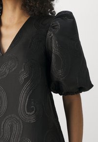 Schwarzes Kleid mit tiefem V-Ausschnitt und Puffärmeln, verziert mit erhabenen, wirbelnden Mustern in glänzender Textur. Glatter Stoff mit dezentem Schimmer.