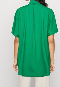 Camicia verde a maniche corte con vestibilità rilassata, caratterizzata da una piega sul retro e una texture liscia. Il colletto è dritto e l'orlo è leggermente arrotondato.