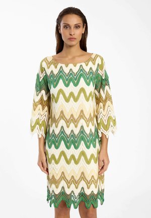 Frau trägt ein knielanges Kleid mit grünen, beigen und weißen Zickzackmustern und dreiviertellangen Ärmeln vor einem schlichten weißen Hintergrund.