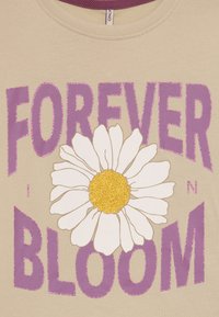 Beige t-skjorte med en stor hvit tusenfryd med en glitrende gul midt. Tekst i lilla som sier "FOREVER IN BLOOM." Glatt bomullsstoff.